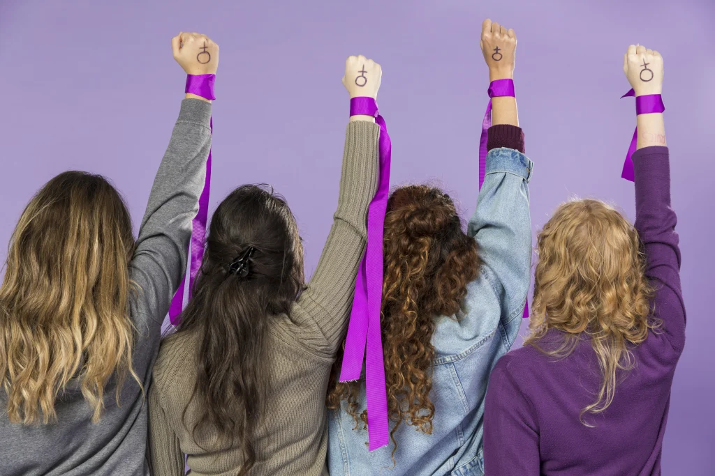 Imagem com quatro mulheres com a mão esquerda levantada com o símbolo das mulheres para simbolizar o combate à desigualdade de gênero