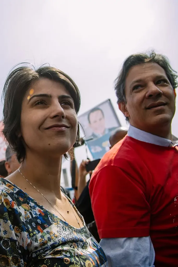 Fotografia de Manuela d’Ávila e Fernando Haddad em um evento de campanha eleitoral ao ar livre. d’Ávila, em primeiro plano, usa uma blusa floral; Haddad, ao seu lado, veste camisa vermelha.