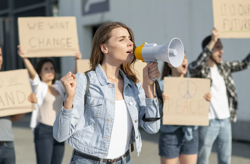 Mulher com um megafone em primeiro plano protestando, com outras quatro pessoas ao fundo seguranado cartazes, pedindo por paz e mudanças. Texto: Lei de paridade de gênero do México.