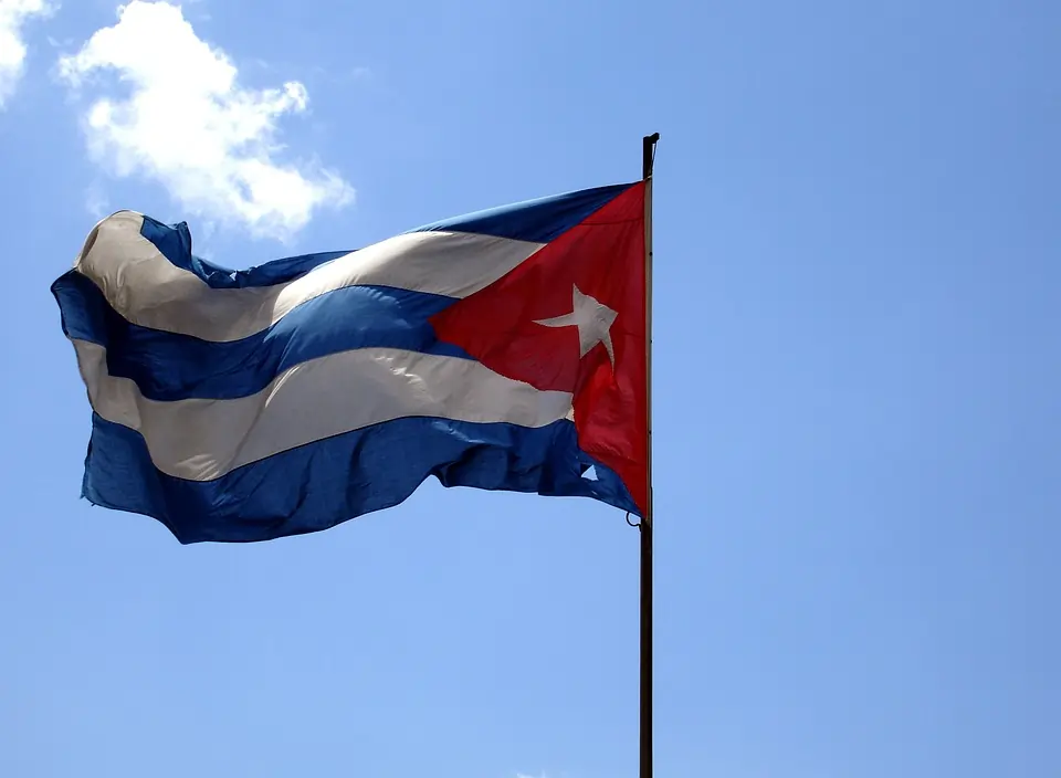A imagem mostra a bandeira de Cuba tremulando contra um céu azul claro e com poucas nuvens. A bandeira, composta por cinco listras horizontais alternadas em azul e branco, possui um triângulo vermelho na lateral esquerda, que contém uma estrela branca de cinco pontas no centro. Texto: A crise dos mísseis de Cuba: os 13 dias à beira da guerra nuclear