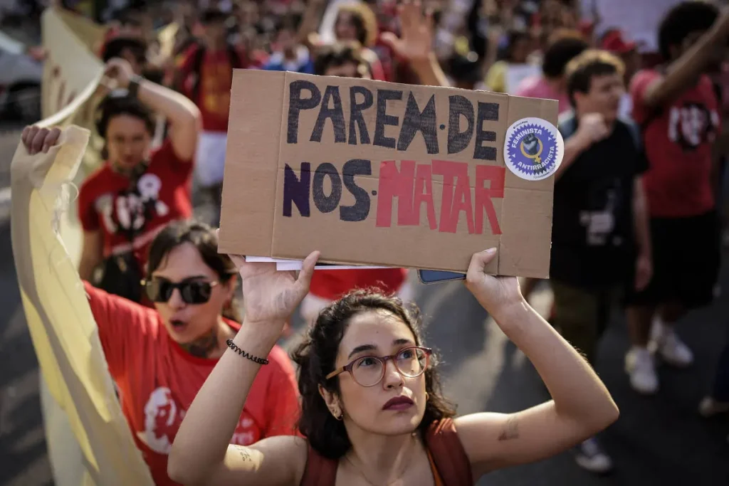 Foto de manifestante com o cartaz “Parem de nos matar” durante marcha pelo Dia Internacional da Mulher na avenida Paulista. Texto: Lei do Feminicídio — 10 anos de avanços e desafios