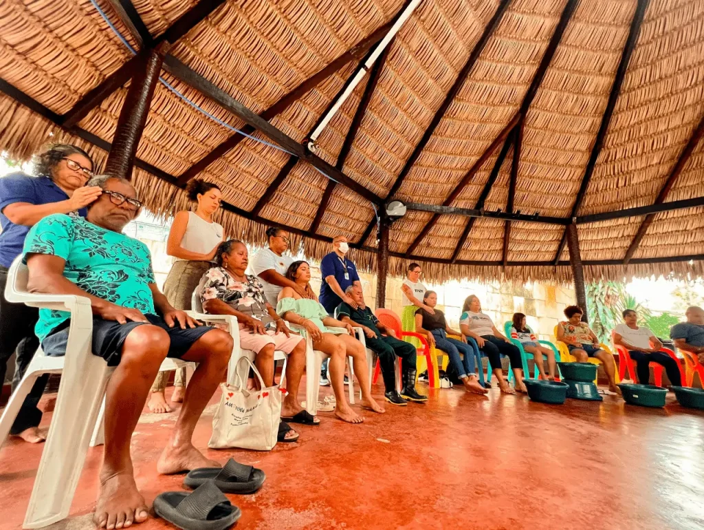 Foto de um grupo de pessoas sentadas em roda, embaixo de uma tenda de palha, durante um encontro sobre entoadas e confluências contracoloniais na saúde mental brasileira. 
