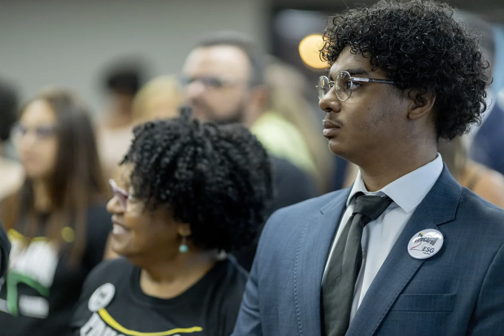 Pessoas em evento institucional, incluindo homem negro de terno e mulher negra com camiseta e broche, representando a diversidade e as carreiras possíveis na Defensoria Pública.