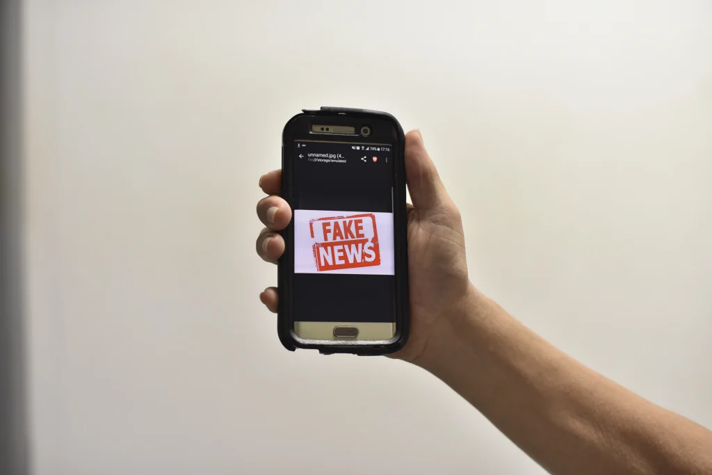 Mão branca segurando um celular com a tela exibindo a mensagem 'Fake News'.