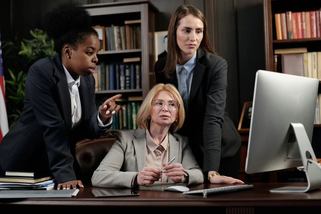 Três mulheres vestindo ternos formais se debruçam sobre uma mesa de escritório, olhando para um monitor de computador. Ao fundo, há estantes com livros, uma bandeira e iluminação ambiente.
