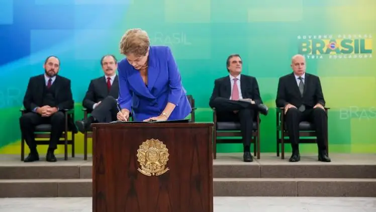 Em cerimônia oficial, a ex-presidenta da República, Dilma Rousseff, assina documento em um púlpito com o brasão da República, em Brasília. Ao fundo, há um painel com as cores da bandeira brasileira e a inscrição "Governo Federal - Brasil".