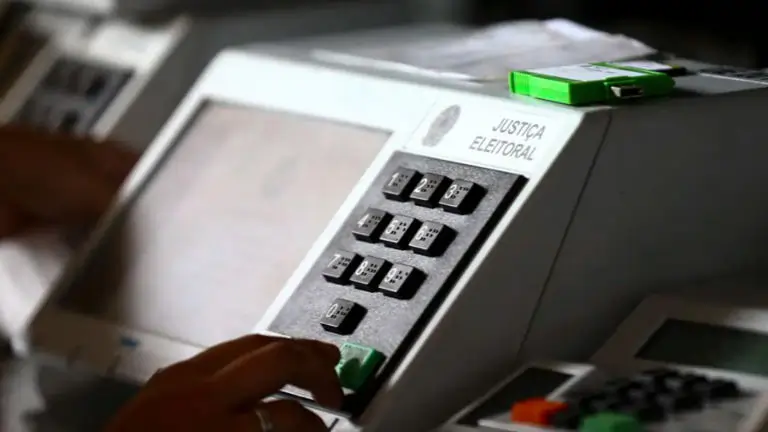 Uma urna eletrônica e uma mão de alguém com o dedo indicador prestes a pressionar a tecla "confirma".