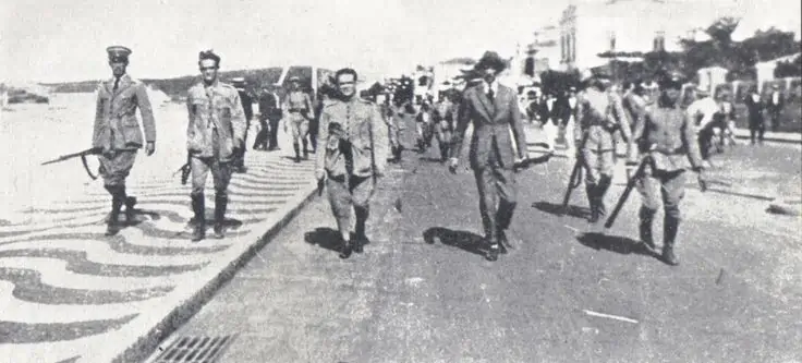 18 homens caminhando na orla de Copacabana, em 1922, pela revolta tenentista, que ocorria no país contra as insatisfações governamentais.