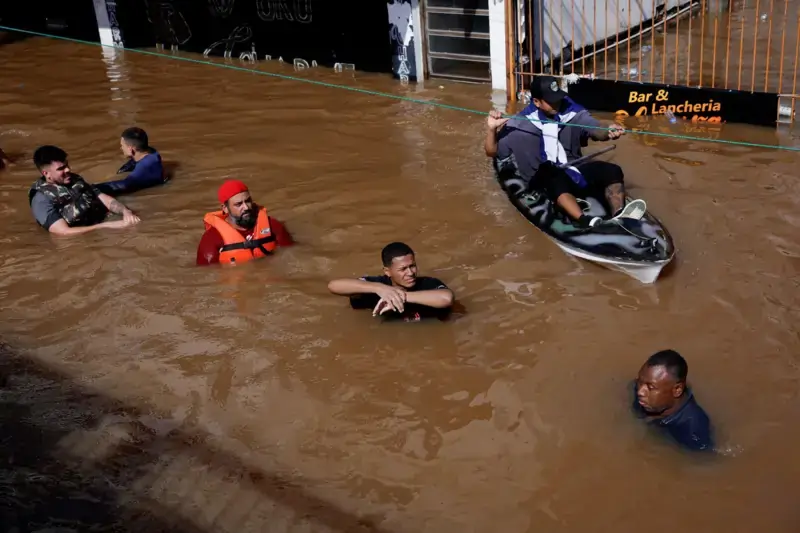 Pessoas atravessam rua alagada durante enchentes no Rio Grande do Sul, com água acima da cintura e uso de bote improvisado para resgate.