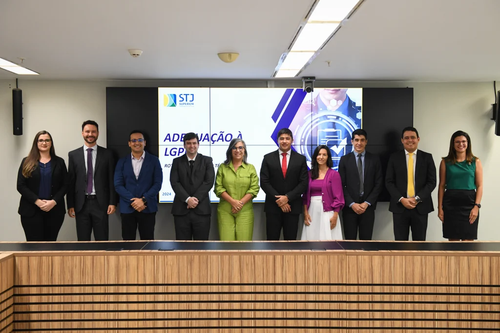 Grupo de homens e mulheres, no total dez pessoas, posicionado lado a lado, de pé em frente ao painel com o título “Adequação à LGPD” destacado.