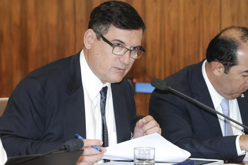 Um homem de cabelos pretos, terno azul escuro e óculos segura um documento enquanto fala em um microfone em reunião de trabalho da Comissão de Juristas da Lei da Arbitragem no Senado Federal.