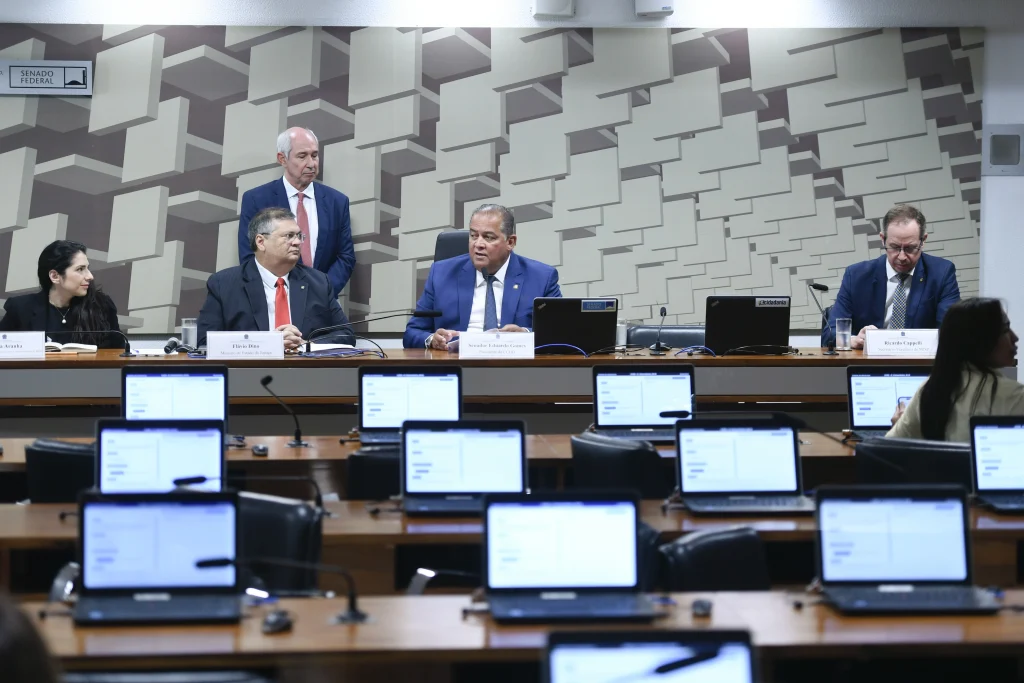 Fotografia em plano médio de uma sala de reuniões no Senado Federal exibindo diversos computadores portáteis abertos e ao fundo, uma mesa de madeira onde encontram-se sentados e conversando três homens e uma mulher, além de outro homem em pé posicionado ao fundo e próximo deles, todos com roupas formais.